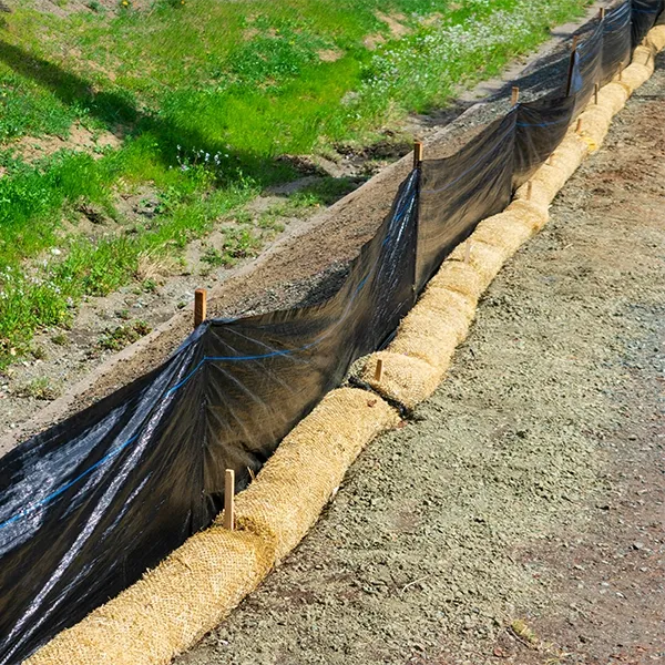 A black plastic sheet is placed over a dirt path next to grass and hay bales for erosion control.
