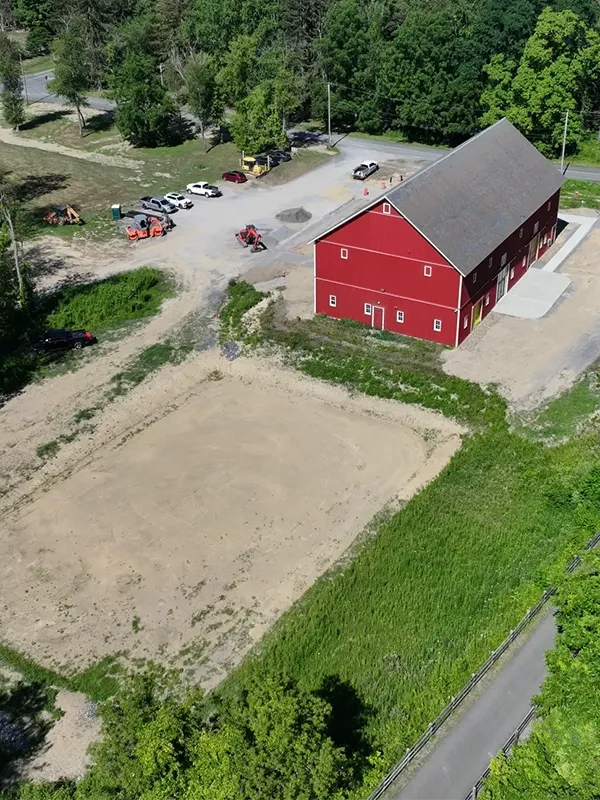 The image is an aerial photograph of a red barn surrounded by vehicles and greenery.